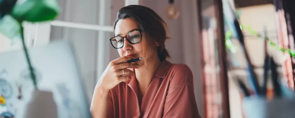 Woman in glasses working on a laptop, holding a pen to her chin and looking thoughtful.