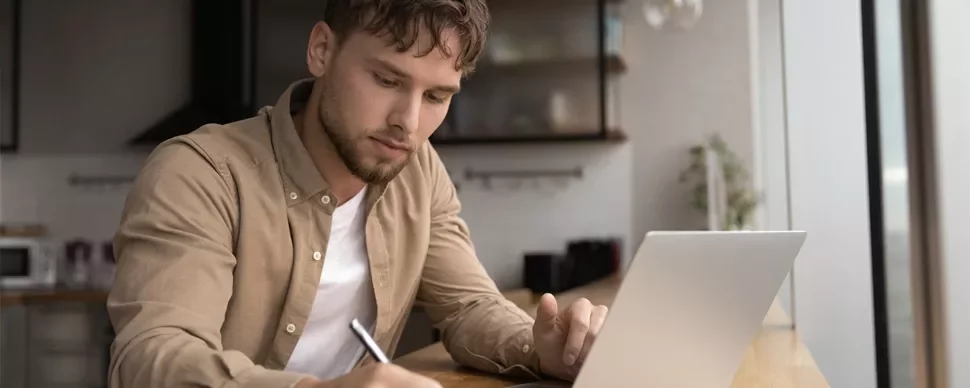 Man writing notes in front of a laptop at a home desk.