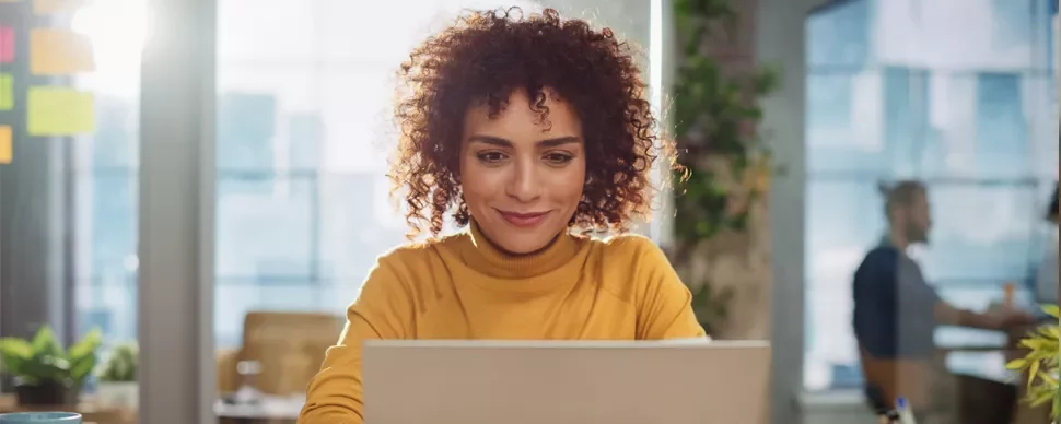 Woman with curly hair working on a laptop in a bright, plant-filled office.