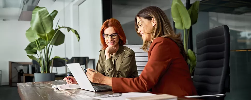 Two women discussing work at a desk in a modern office with large plants.