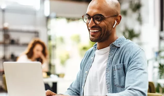 Smiling man wearing wireless earbuds, working on a laptop.