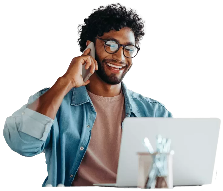 A person seated at a desk using a laptop, holding a phone to their ear, with a small desk accessory in front.