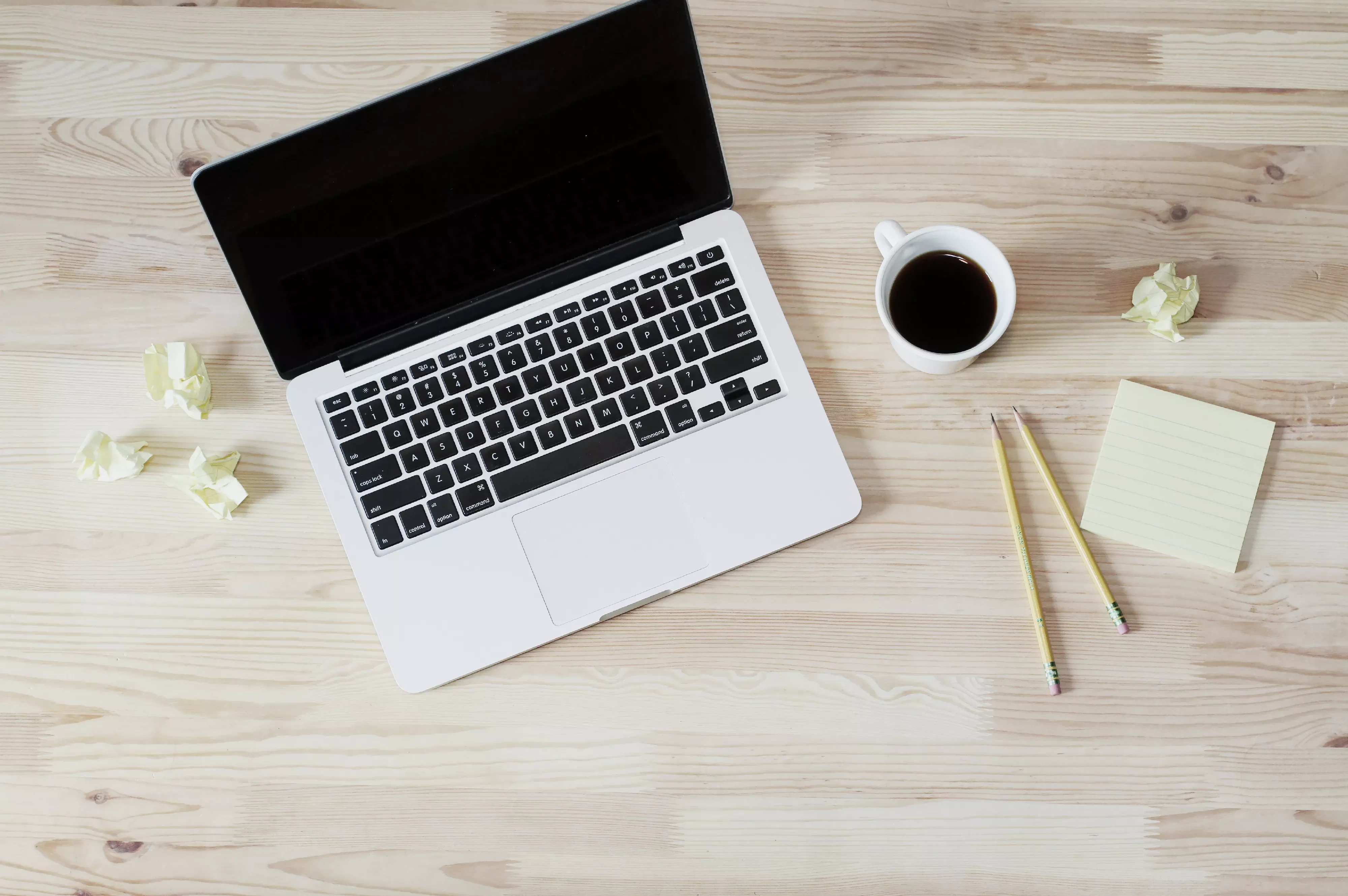 Open laptop on wooden desk with coffee, pencils, sticky notes, and crumpled paper.