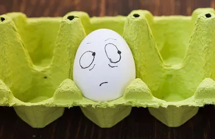 Egg with a drawn face showing confusion in a green egg carton.