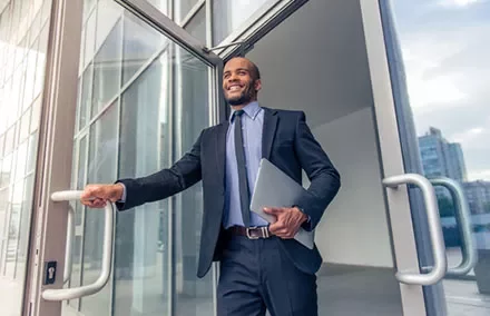 Businessman in a suit exiting a glass building, holding a laptop and smiling.