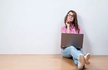 Woman sitting, thinking with a laptop on her lap against a white wall.
