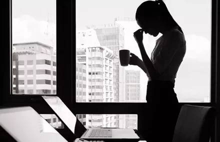 Silhouette of a woman holding a mug, standing by a window, overlooking city buildings, with laptops in the foreground.