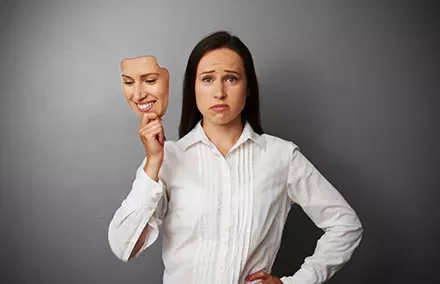 Woman holding a mask of a smiling face, revealing her own sad expression.