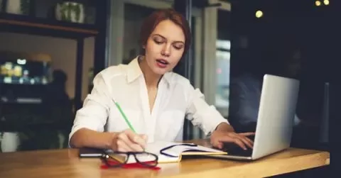 Woman writing notes and using a laptop at a cafe.