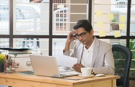 Man at desk reading paper by laptop with post-it notes on the window.
