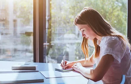 Woman writing at a sunlit desk.
