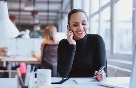 Smiling woman on phone at desk, taking notes in a bright office.