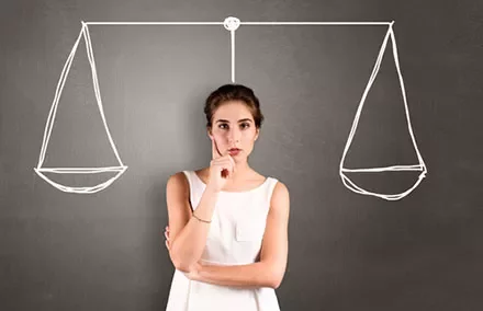 Woman pondering, standing in front of sketched balance scales on a blackboard.