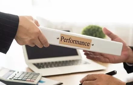 Two people exchanging a binder labeled 'Performance' at a desk with a laptop and calculator.