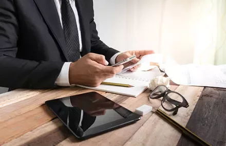 Businessman in suit using smartphone at a desk with tablet and glasses.