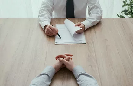 Two people sitting at a table, one holding a pen and paper, in an office setting.