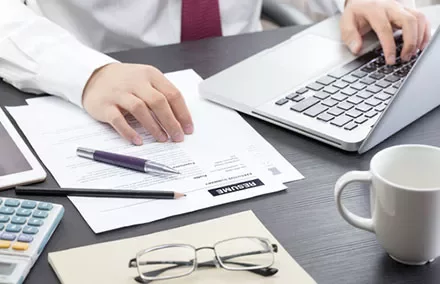 Man in office working on documents and laptop with a calculator, mug, and glasses on the desk.