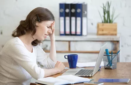 Woman holding head in frustration at desk with laptop and coffee.