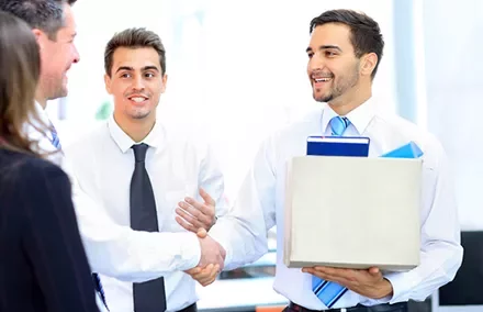 A man holding a box is smiling and shaking hands with another man in an office setting.