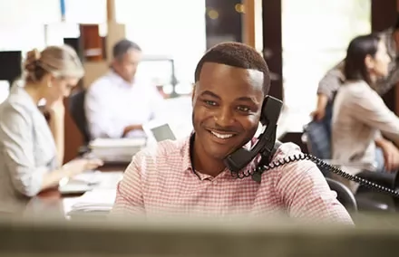 Man smiling while talking on the phone in a busy office.