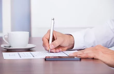 Person signing a paper at a desk with a pen and smartphone nearby.