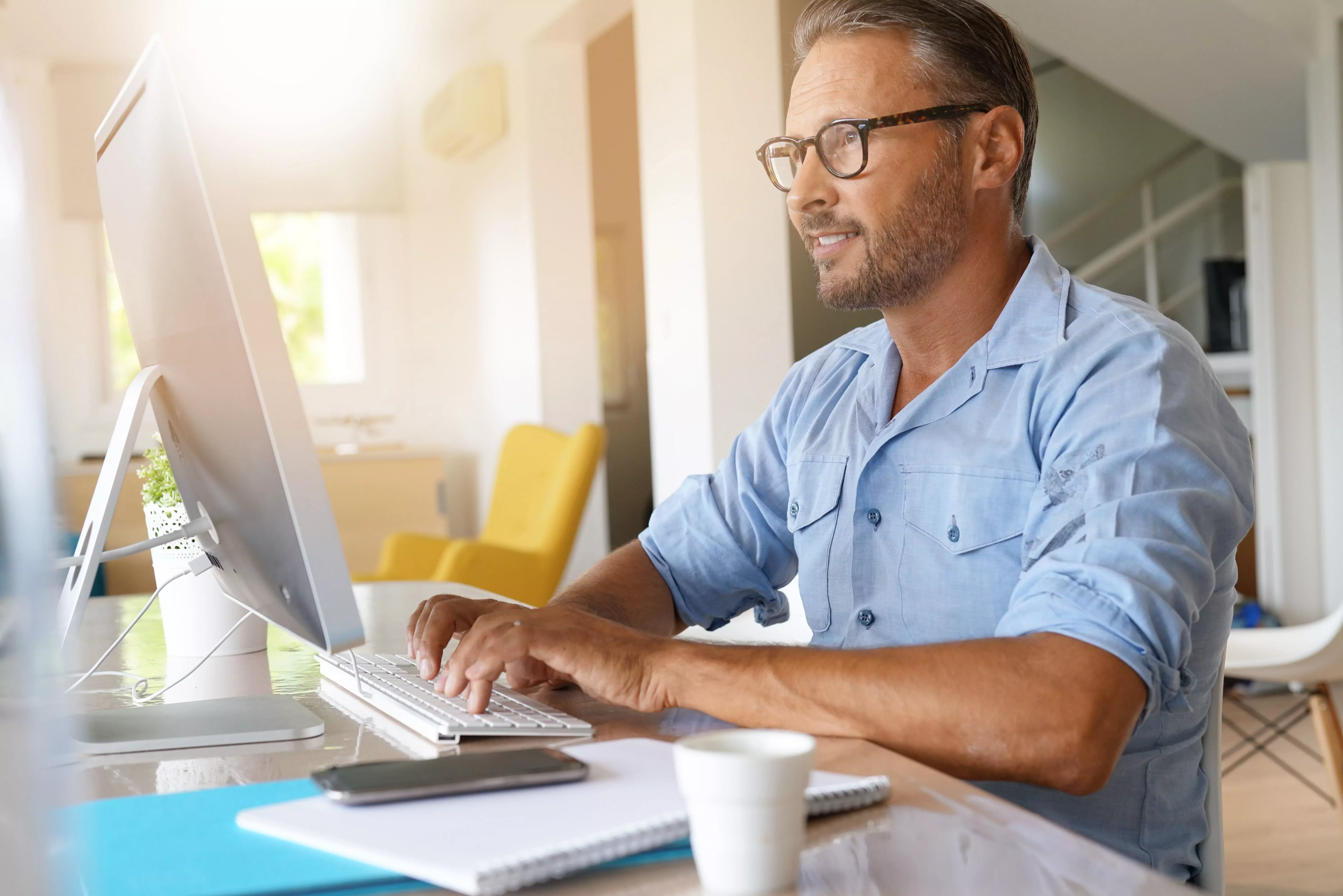 Man in glasses working at a desktop computer in a bright room.