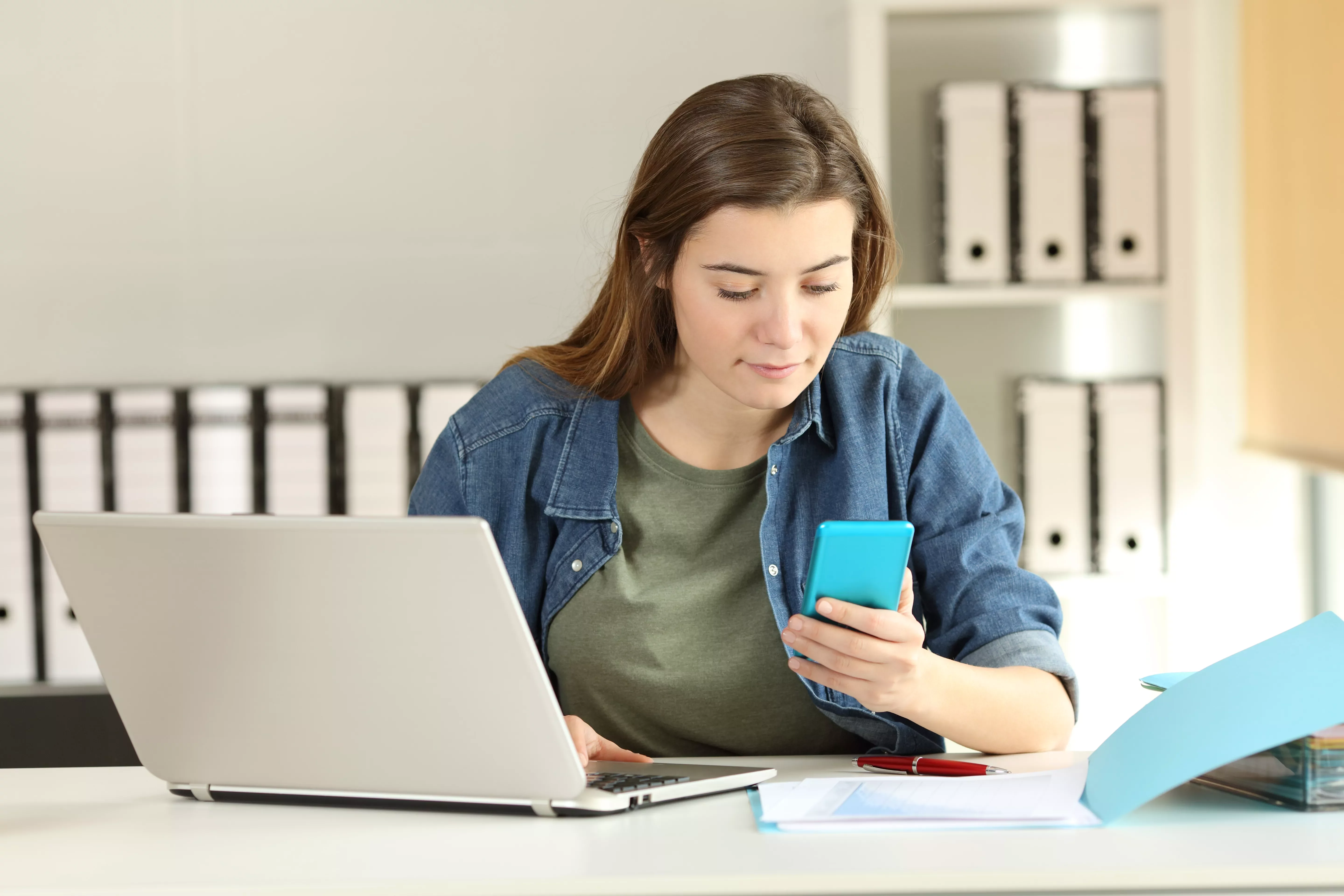 Woman in denim shirt checks phone at desk with laptop.