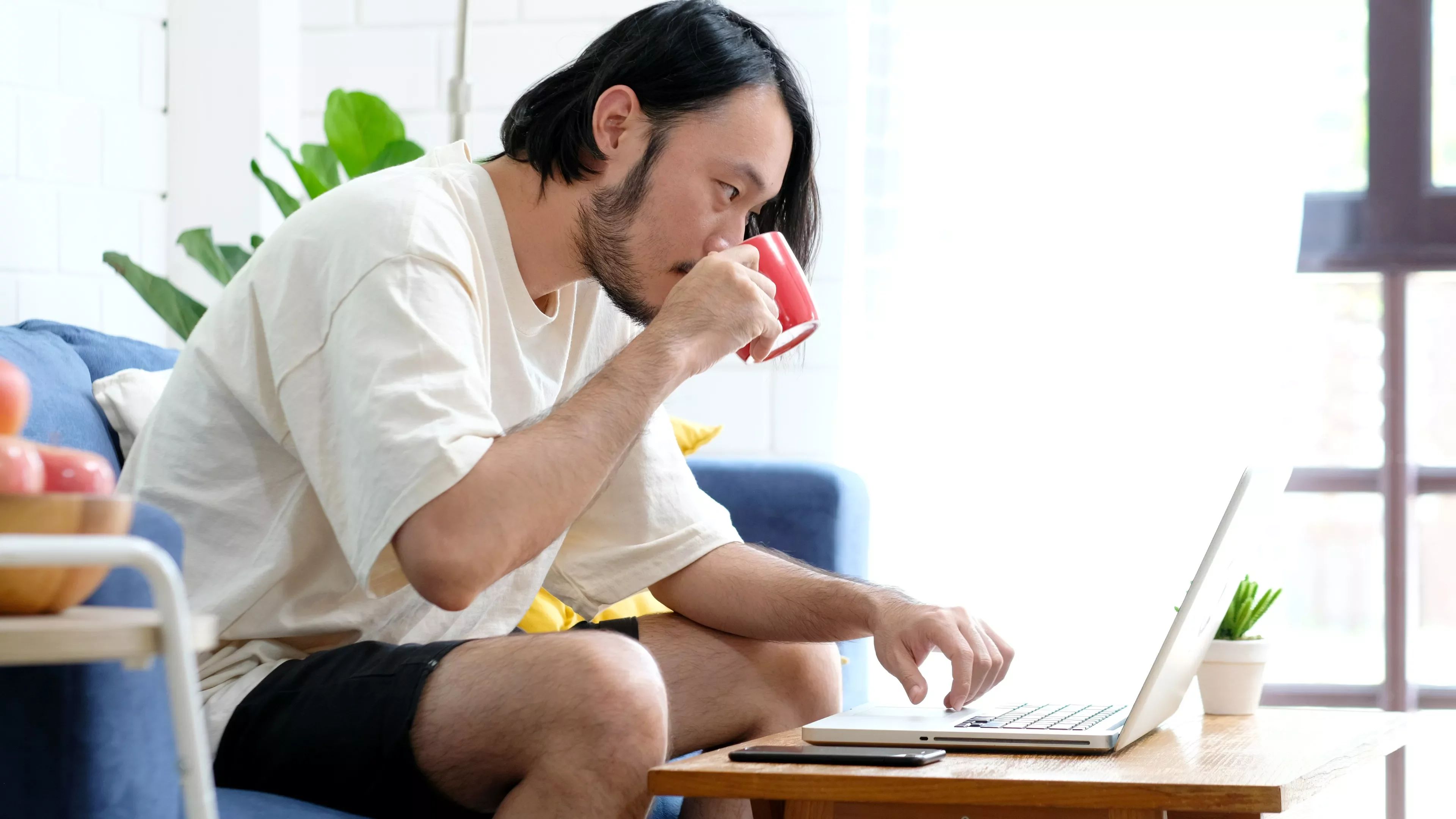 Man in a white shirt drinking coffee while working on a laptop.
