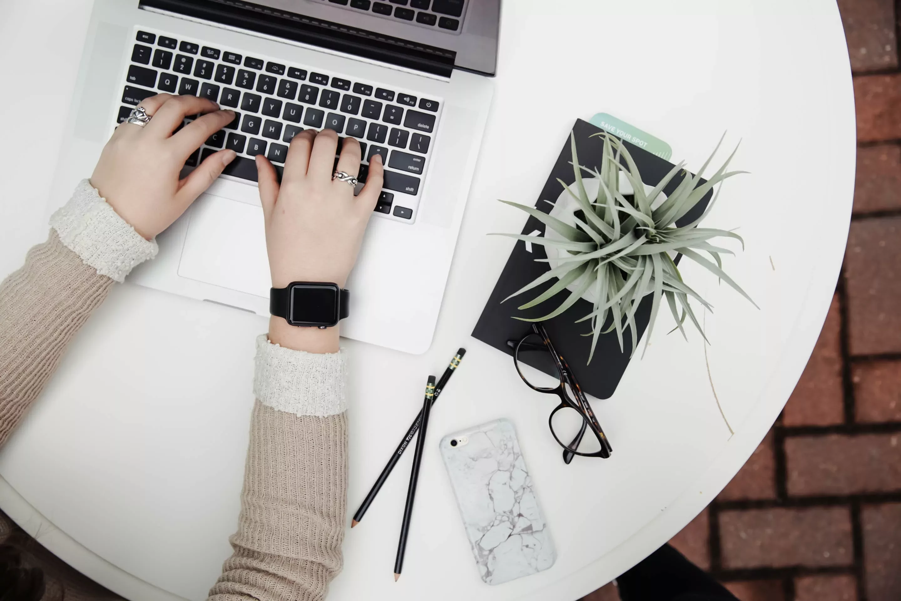Hands typing on a laptop beside a plant and glasses on a white table.