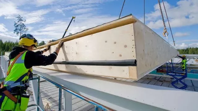 Construction worker guides a large wooden panel into place at a building site under a clear sky.