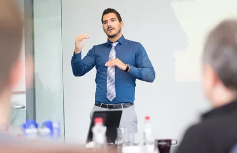 Man giving a presentation in a blue shirt and tie, gesturing with hands.