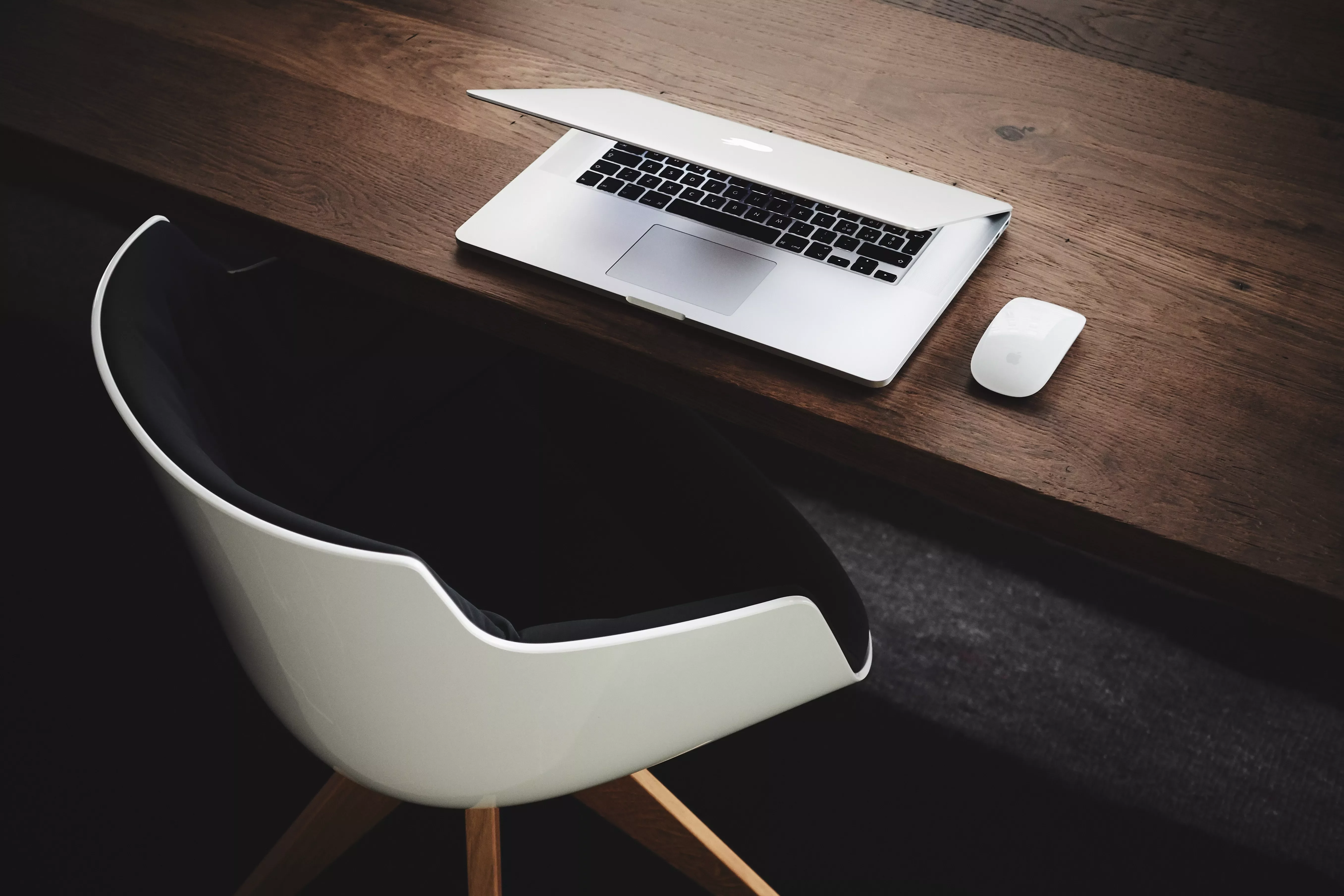 Open laptop and mouse on a wooden desk with a modern chair.