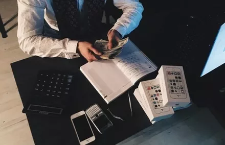 Person counting money at a desk with phones and a notebook.