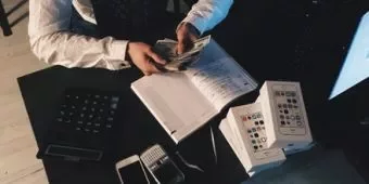 Person counting money at desk with calculator and phone boxes.