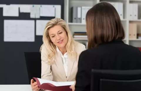 A woman in a beige blazer discusses a document with another woman in an office.
