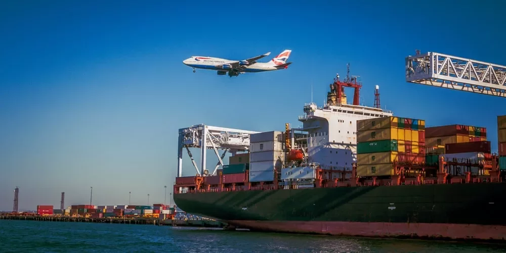 A cargo ship in port with colorful containers and an airplane flying overhead under a clear blue sky.