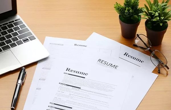 Laptop, resumes, pen, glasses, and potted plants on a wooden desk.