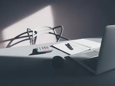 A laptop, notepad, pen, and calculator on a sunlit desk.