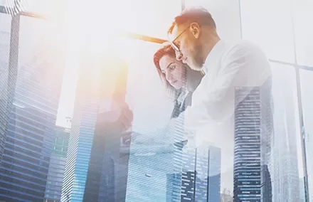 Double exposure of two business professionals working on a tablet with skyscrapers and sunlight in the background.