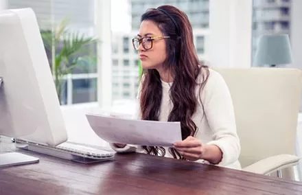 Woman in glasses working at a computer, holding documents.