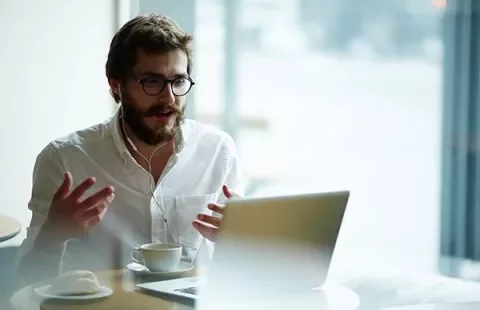 Man wearing headphones, engaged in a video call on a laptop, sitting at a cafe with a cup of coffee.