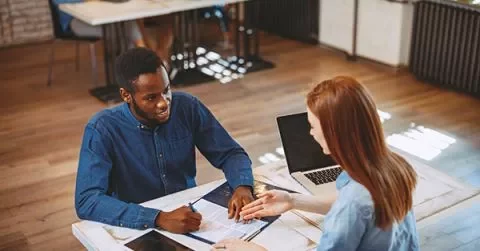 Two people discussing over documents at a table with laptops in a bright office.